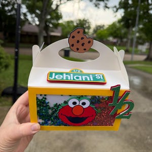 May include: A white and yellow box with a clear window, featuring an Elmo design and the text "Jehani St". The box is decorated with a cookie and the number "1/2". The box is filled with colorful confetti.