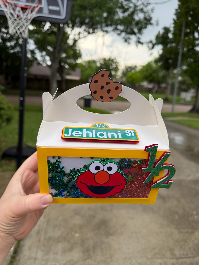 May include: A white and yellow box with a clear window, featuring an Elmo design and the text "Jehani St". The box is decorated with a cookie and the number "1/2". The box is filled with colorful confetti.