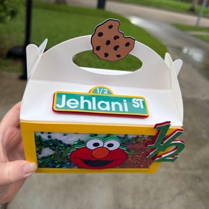 May include: A white and yellow party favor box with a handle, featuring a cutout cookie, the text "Jehlanl ST", and an Elmo graphic. The box is decorated with green and red accents and the number "1/2".
