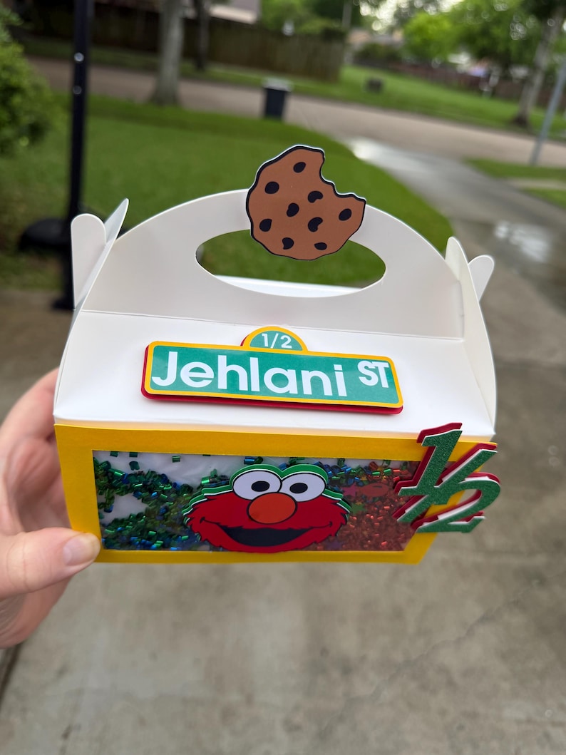 May include: A white and yellow party favor box with a handle, featuring a cutout cookie, the text "Jehlanl ST", and an Elmo graphic. The box is decorated with green and red accents and the number "1/2".