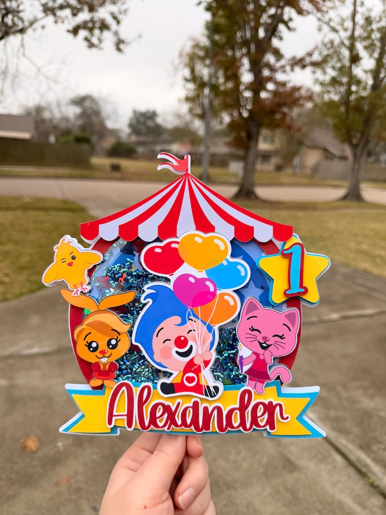 May include: A colorful birthday cake topper featuring a circus tent design with red and white stripes. The topper includes cartoon characters, balloons, and the name "Alexander" in red script. A large number "1" is also included.