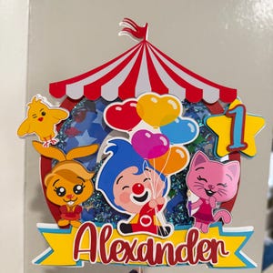 Puede incluir: Un adorno para pastel colorido con un dise&ntilde;o de carpa de circo a rayas rojas y blancas. El adorno incluye personajes de dibujos animados, globos y el nombre "Alexander" en escritura roja. Tambi&eacute;n se incluye una estrella amarilla con el n&uacute;mero "1".