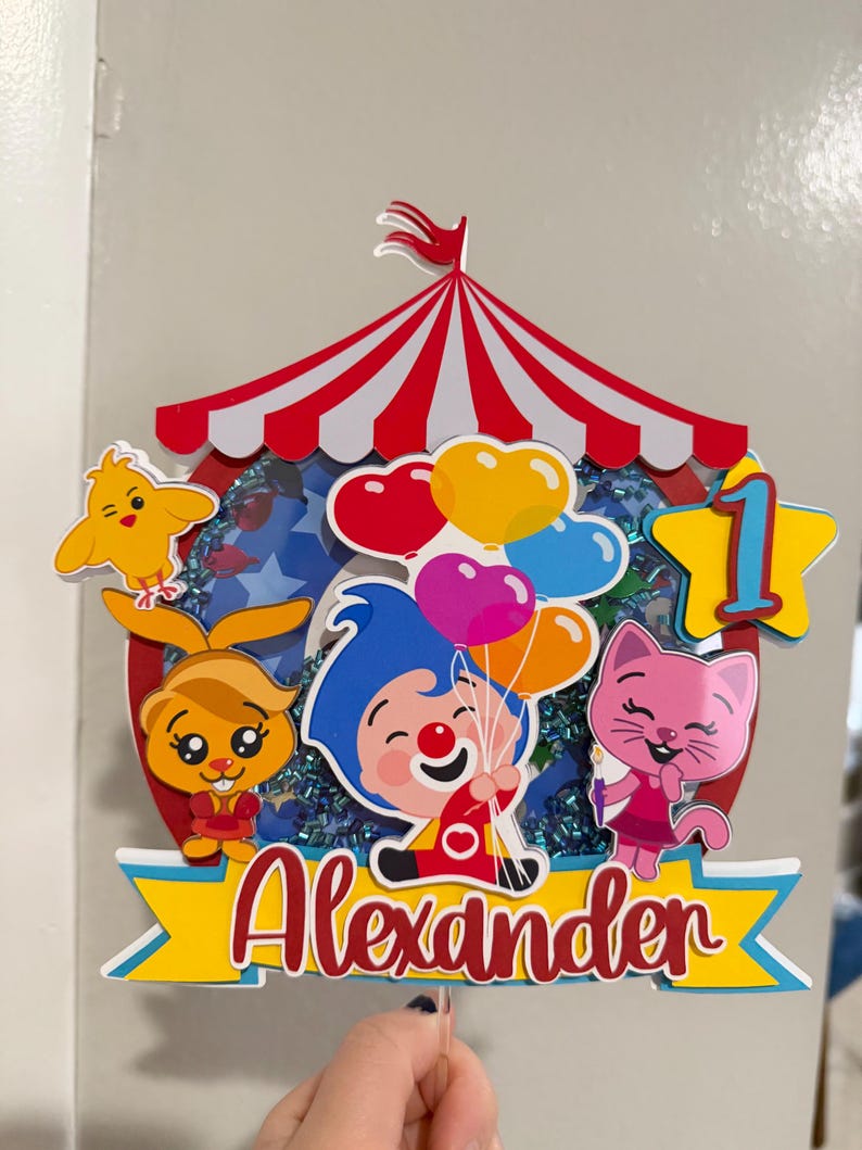 Puede incluir: Un adorno para pastel colorido con un dise&ntilde;o de carpa de circo a rayas rojas y blancas. El adorno incluye personajes de dibujos animados, globos y el nombre "Alexander" en escritura roja. Tambi&eacute;n se incluye una estrella amarilla con el n&uacute;mero "1".