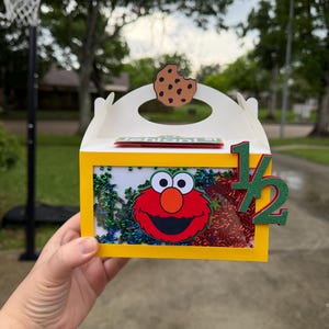 May include: A yellow box with a clear window featuring Elmo's face and colorful confetti. A brown cookie cutout sits on top. The number "1/2" is attached to the side. The box is held by a hand.