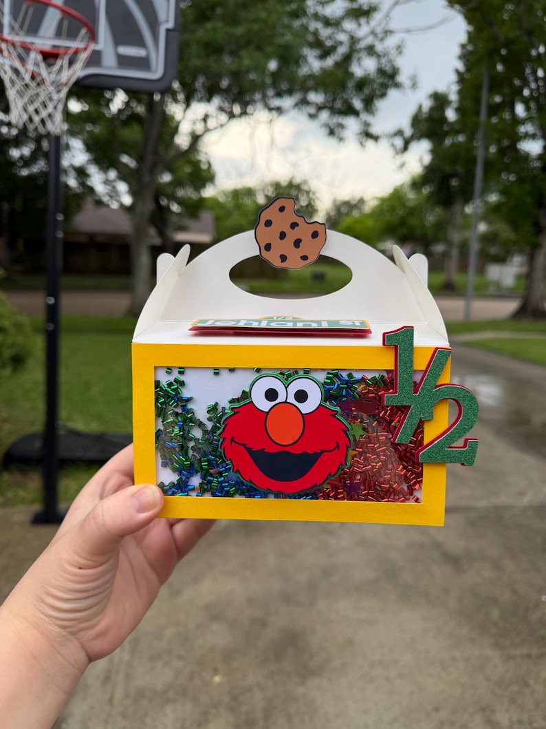 May include: A yellow box with a clear window featuring Elmo's face and colorful confetti. A brown cookie cutout sits on top. The number "1/2" is attached to the side. The box is held by a hand.