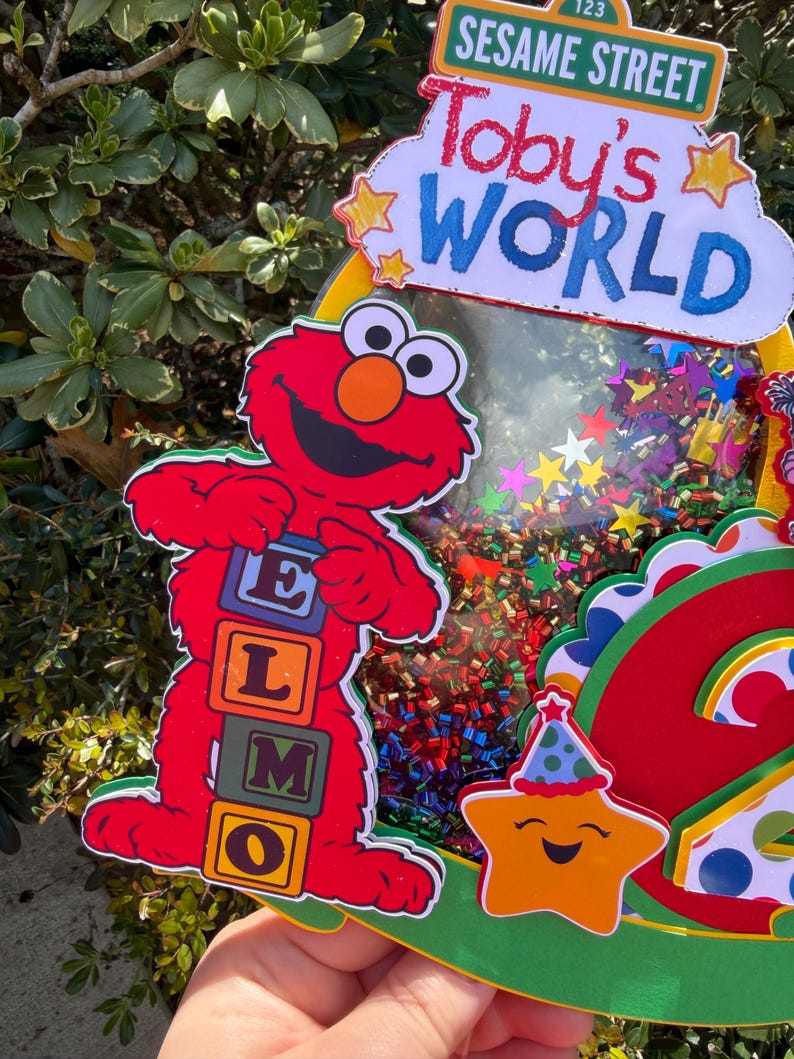 May include: A colorful Sesame Street-themed decoration featuring Elmo holding letter blocks. The decoration includes the text "Toby's WORLD" and a smiling star with a party hat. The background is filled with confetti.