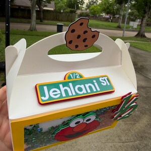 May include: A white cardboard box with a yellow trim, featuring a cutout cookie on top and an Elmo character design. The box has the text "1/2 Jehlani ST" on it.