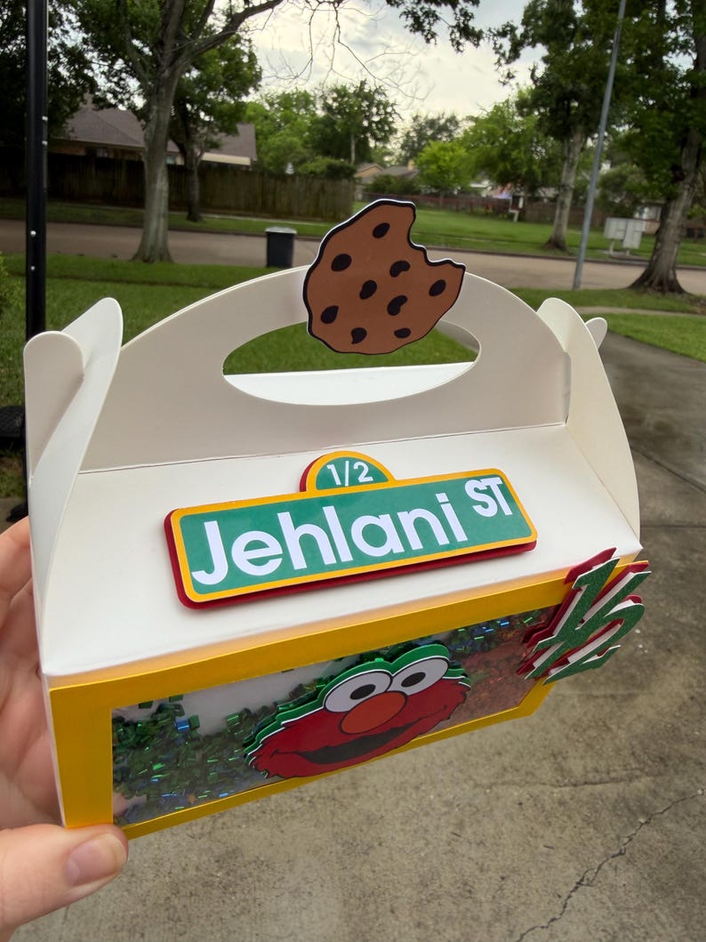 May include: A white cardboard box with a yellow trim, featuring a cutout cookie on top and an Elmo character design. The box has the text "1/2 Jehlani ST" on it.