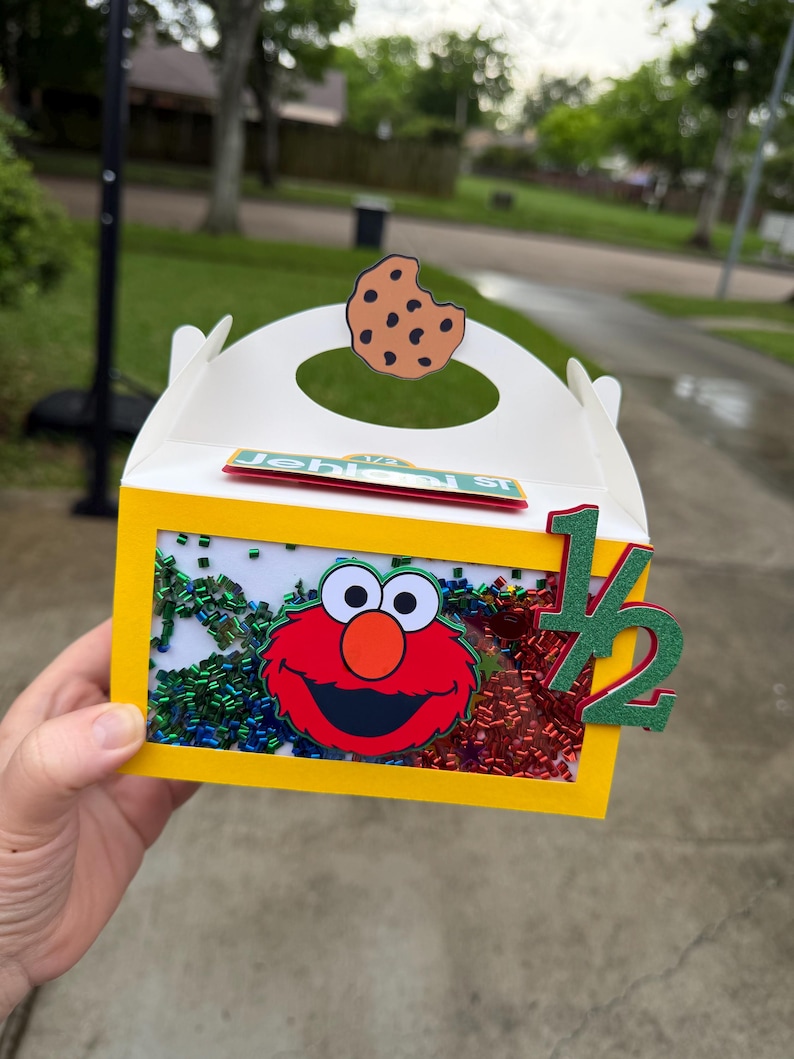 May include: A yellow and white party favor box featuring Elmo from Sesame Street. The box has a clear window filled with colorful confetti, a cookie cutout, and the number "1/2" in green. The box is decorated with the name "Jehlemi St."