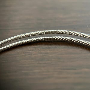 May include: Close-up of two silver-colored, twisted metal bangles. The bangles are thin and have a rope-like texture. The background is a dark brown wooden surface. The bangles have markings on them.