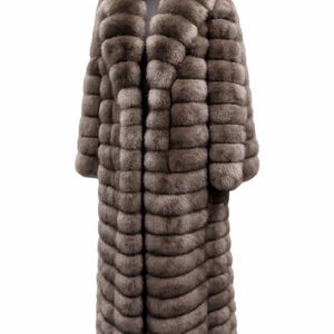 May include: Full-length, gray fur coat with a wide collar and long sleeves. The coat is displayed on a hanger against a white background. The fur appears soft and has a horizontal, ribbed design.