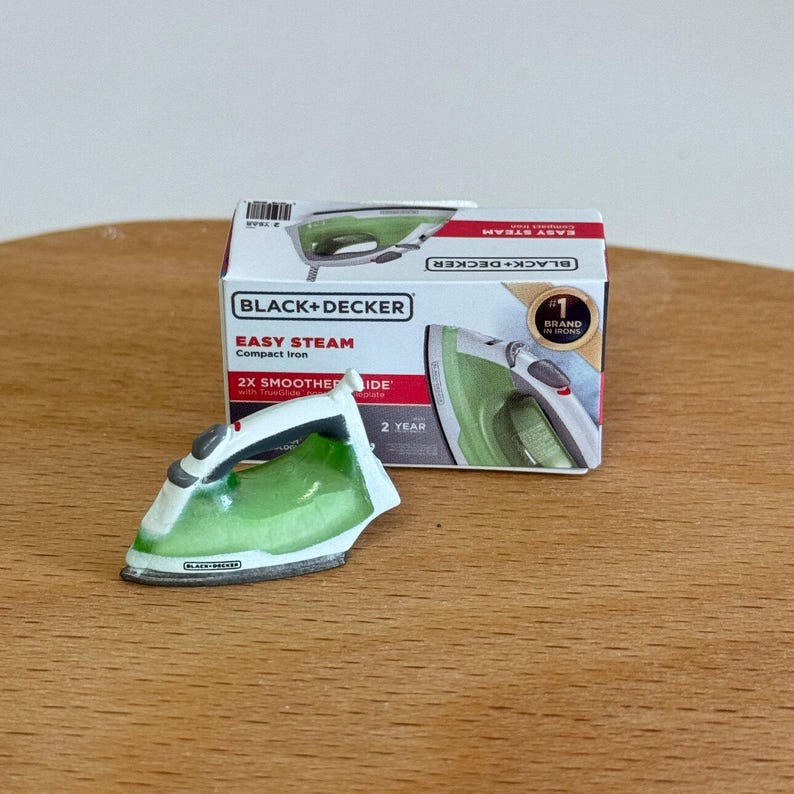 以下が含まれることがあります： 緑と白のデザインの、Black+Decker Easy Steamコンパクトアイロンのミニチュア。アイロンは、アイロンと「2X Smoother Glide」の文字も表示されている製品ボックスの隣にあります。
