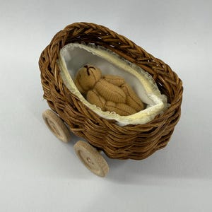 Puede incluir: Un cochecito de bebé en miniatura de mimbre marrón con ruedas de madera. En el interior, un pequeño osito de peluche beige descansa sobre una cama con adornos blancos y amarillos. El cochecito está diseñado para una muñeca u otro juguete pequeño.