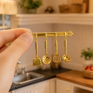May include: A miniature gold-colored kitchen utensil rack featuring four hanging tools: a flat spatula, a perforated skimmer, a slotted turner, and a small ladle. The metallic rack has a horizontal bar with four hooks for the utensils.