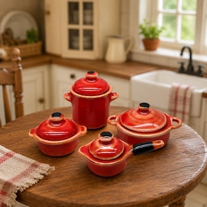 Puede incluir: Un juego de cuatro piezas de cocina en miniatura de cerámica roja, que incluye un bote alto, dos ollas redondas pequeñas con asas laterales y un cazo pequeño con un mango negro largo, todos con tapas a juego y pomos de color negro.