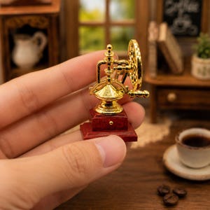May include: A miniature coffee grinder featuring a shiny gold-toned metal top mechanism with a side crank handle and a circular wheel, mounted on a small, square, dark reddish-brown wooden base with a tiny gold knob on the front drawer.