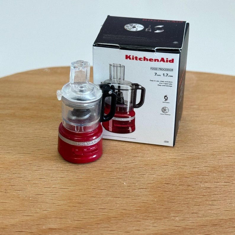 以下が含まれることがあります： 透明な蓋と黒いハンドルが付いた赤いKitchenAidフードプロセッサー。製品は、KitchenAidのロゴと製品の詳細が記載された元のパッケージの横に表示されています。