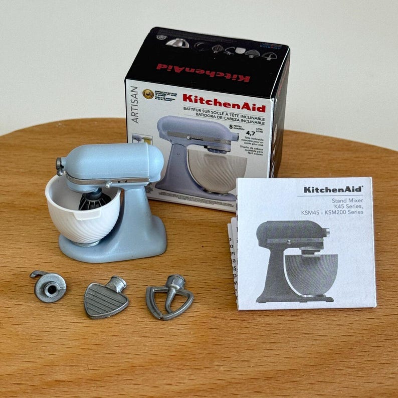 以下が含まれることがあります： ライトブルーのミニチュアKitchenAid Artisanスタンドミキサー。白いボウルと3つのアタッチメントが付いています。ミキサーは箱と小さな取扱説明書と一緒に表示されています。箱にはKitchenAidのロゴと「Artisan」と「Batteur sur socle &agrave; t&ecirc;te inclinable」の文字があります。