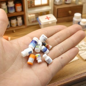 Pode incluir: Uma coleção de frascos e recipientes de medicamentos em miniatura de plástico em várias formas e cores, incluindo branco, laranja e azul, com etiquetas impressas com texto, agrupados para representar um conjunto de farmácia em pequena escala.