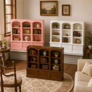 Op de afbeelding: Miniatuur boekenkasten in roze, wit en donkere houttinten. Elke plank is gevuld met boeken, decoratieve items en kleine planten. De scène speelt zich af in een kamer met een patroonkleed en meubels in antieke stijl.