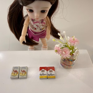 May include: A doll wearing a pink outfit with a Hello Kitty graphic stands near miniature cigarette packs. Two packs of Camel Filters and two packs of Marlboro cigarettes are visible. A small vase with pink flowers is also present.