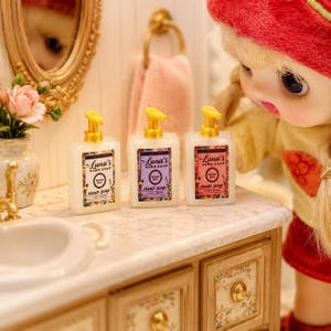 May include: Three miniature rectangular hand soap bottles with gold pump dispensers, each featuring a label that reads Luna's Hand Soap. The bottles are white, purple, and pink, arranged on a marble-patterned countertop next to a small vase.