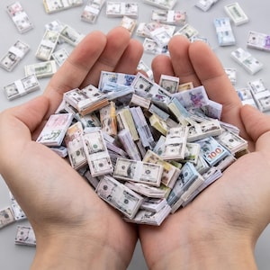 May include: A collection of miniature stacks of United States dollar bills, featuring various denominations with detailed printing, held together in a cupped arrangement, with additional loose miniature stacks scattered across the surrounding area.