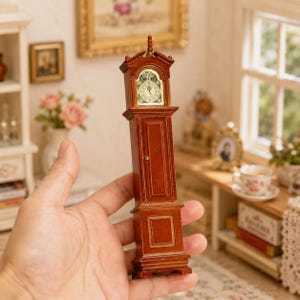Pode incluir: Um relógio de pé em miniatura de madeira com um acabamento castanho rico, apresentando um topo arqueado clássico, uma porta central com um pequeno puxador de latão e um mostrador branco com numerais romanos e ponteiros pretos.