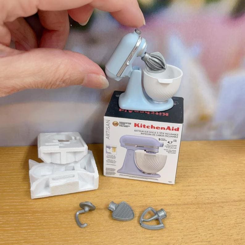 以下が含まれることがあります： 白いボウルと泡立て器の付属品が付いた、水色のミニチュアKitchenAidスタンドミキサー。ミキサーは、KitchenAidのロゴと製品の詳細が記載された小さな箱の上にあります。追加のミキサーアタッチメントと白いプラスチックコンポーネントも存在します。