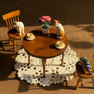 May include: A miniature wooden dining set featuring a round table and two spindle-back chairs, arranged on a white crocheted doily. The table holds two small teacups with saucers, a potted plant with pink flowers, and three small cat figurines.