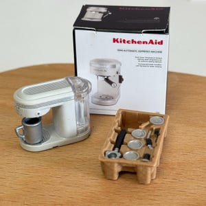 以下が含まれることがあります： 白いミニチュアのKitchenAidセミオートエスプレッソマシンと銀色のカップ。後ろの箱にはマシンとアクセサリーが表示されています。茶色のトレイには追加のエスプレッソマシンの部品が入っています。