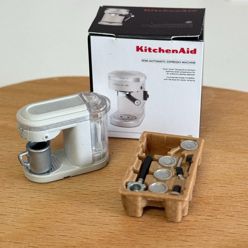 以下が含まれることがあります： 白いミニチュアのKitchenAidセミオートエスプレッソマシンと銀色のカップ。後ろの箱にはマシンとアクセサリーが表示されています。茶色のトレイには追加のエスプレッソマシンの部品が入っています。
