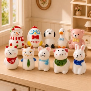 May include: A collection of ceramic figurines in the shape of animals and a snowman. The figurines are white with colorful accents, including a red scarf, blue bow tie, and green scarf. The collection includes a dog, duck, bear, and rabbit.