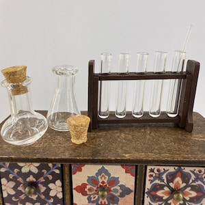 May include: Miniature glass laboratory equipment set, including a flask with a cork, a beaker, a test tube rack with test tubes, and a stirring rod. The set is displayed on a wooden surface with decorative drawers below.