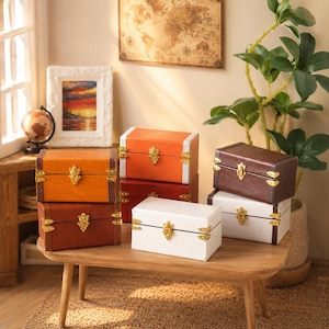 May include: A collection of six rectangular storage chests in various colours including orange, red, brown, and white. Each chest features a hinged lid, gold-toned metal corner reinforcements, and a central gold-toned latch closure mechanism.