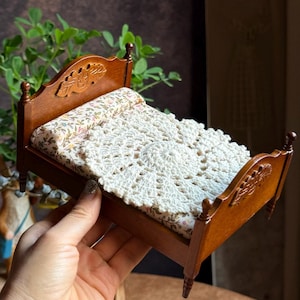 May include: A miniature wooden bed frame with a dark brown finish, featuring carved headboard and footboard details. The bed is dressed with a floral patterned mattress and a round, white, crocheted lace doily placed on top as a coverlet.