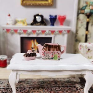 May include: A miniature pink ceramic teapot and lid, designed to resemble a cottage, sit on a white table. The teapot has a brown thatched roof and is decorated with red berries and a green door. The scene suggests a dollhouse or miniature setting.