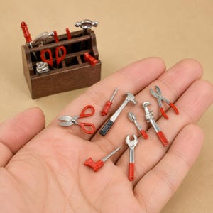 Op de afbeelding: Een miniatuur houten gereedschapskist met diverse kleine metalen gereedschappen met rode handvatten, waaronder hamers, tangen en moersleutels. Extra miniatuurgereedschap met rode en zilveren accenten wordt in de handpalm getoond.