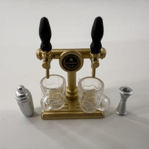 Puede incluir: Un grifo de cerveza en miniatura con dos tazas transparentes, una coctelera plateada y un jigger plateado. El grifo de cerveza es de color dorado con asas negras y el texto "SUPER BEER".