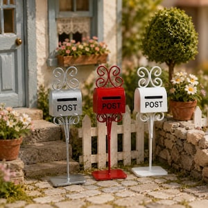 Può includere: Miniature cassette postali in argento, rosso e bianco, ciascuna con un design a volute decorativo e la parola "POST". Le cassette postali sono su piedistalli davanti a una staccionata bianca e un muro di pietra, con fiori e una casa sullo sfondo.