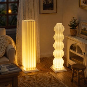 Può includere: Due lampade da terra illuminate. Una è una colonna alta, bianca e scanalata. L'altra è una lampada bianca a forma di onda. Entrambe le lampade sono su basi quadrate bianche ed emettono una calda luce.