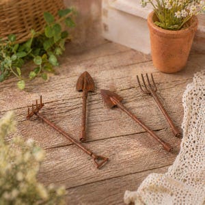 Pode incluir: Quatro mini ferramentas de jardim castanho enferrujado: um ancinho, duas pás e um forcado. As ferramentas estão dispostas sobre uma superfície de madeira, com um pequeno vaso de flores de terracota no fundo. As ferramentas são provavelmente para um jardim em miniatura.