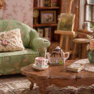 Può includere: Scena in miniatura di soggiorno per casa delle bambole con un divano verde floreale, un tavolino di legno e un servizio da tè. Il servizio da tè comprende una teiera, una tazza e un piattino con motivi floreali. Un cuscino decorativo e un piccolo libro sono anche sul tavolo.