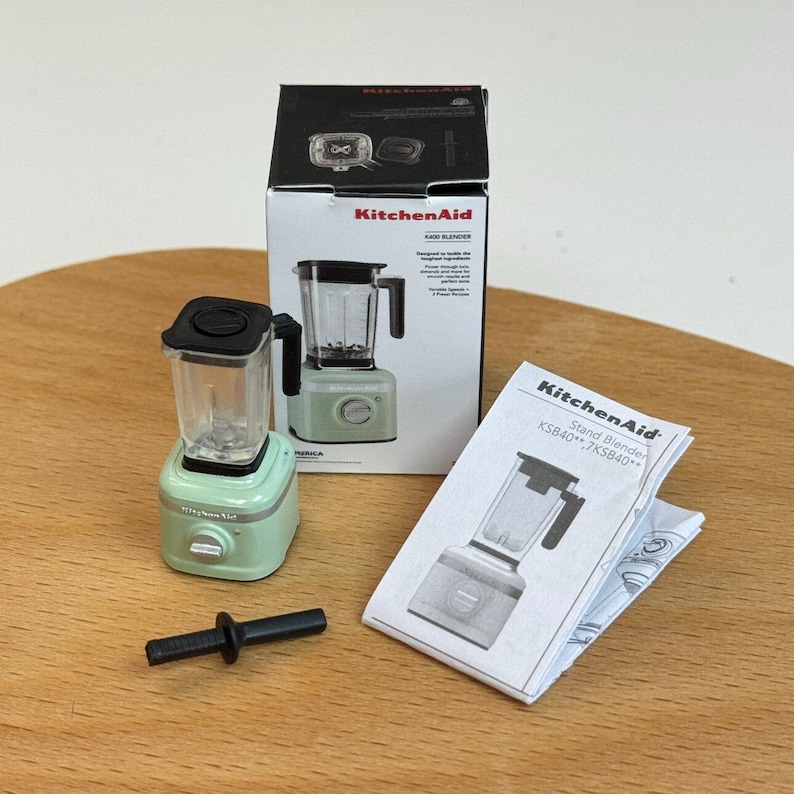 以下が含まれることがあります： 黒いピッチャーとハンドルが付いた、ミントグリーンのミニチュアKitchenAidスタンドブレンダー。ブレンダーは、箱、小さな取扱説明書、黒い混合アタッチメントと一緒に表示されています。箱には「KitchenAid」と「5000 Blender」と書かれています。