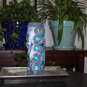 May include: A decorative, cylindrical vase with a textured, gray surface and turquoise glass accents. The vase is displayed on a silver tray, with potted plants in the background. The vase is approximately 8 inches tall.