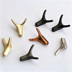 Puede incluir: Una colección de ganchos decorativos con forma de cuerno en varios acabados metálicos, incluyendo dorado, bronce, negro y plateado. Estos ganchos están diseñados para montaje en pared y ofrecen una estética rústica pero moderna. Los ganchos se muestran sobre un fondo blanco.