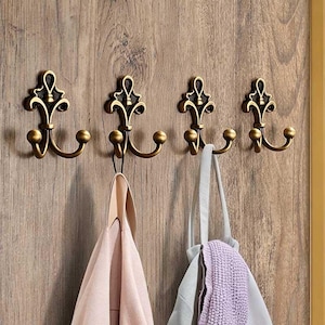 Peut inclure: Quatre patères murales en métal couleur laiton antique avec une forme décorative inspirée de la fleur de lys et deux bras incurvés. Chaque crochet présente un montant central vertical avec un embout arrondi et deux crochets latéraux.