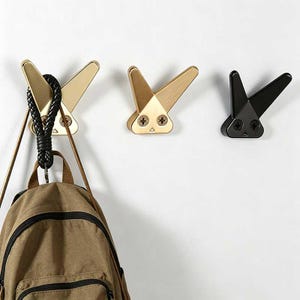 Puede incluir: Tres perchas de pared decorativas con forma de conejo, doradas, bronce y negras. Una mochila cuelga de la percha dorada. Las perchas están fijadas a una pared blanca.