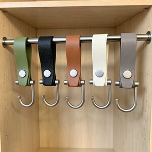 May include: Five leather-like hanging hooks suspended from a horizontal metal rod. Each hook features a folded strap in olive green, black, tan, cream, or grey, secured with a silver-toned metal snap button and a matching silver-toned metal hook.