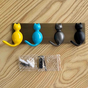 Puede incluir: Un perchero de pared con cuatro ganchos en forma de gato en colores amarillo, azul, gris oscuro y negro, fijados a una placa metálica rectangular de tono bronce. Incluye dos bolsas de plástico con tornillos y tacos para el montaje.